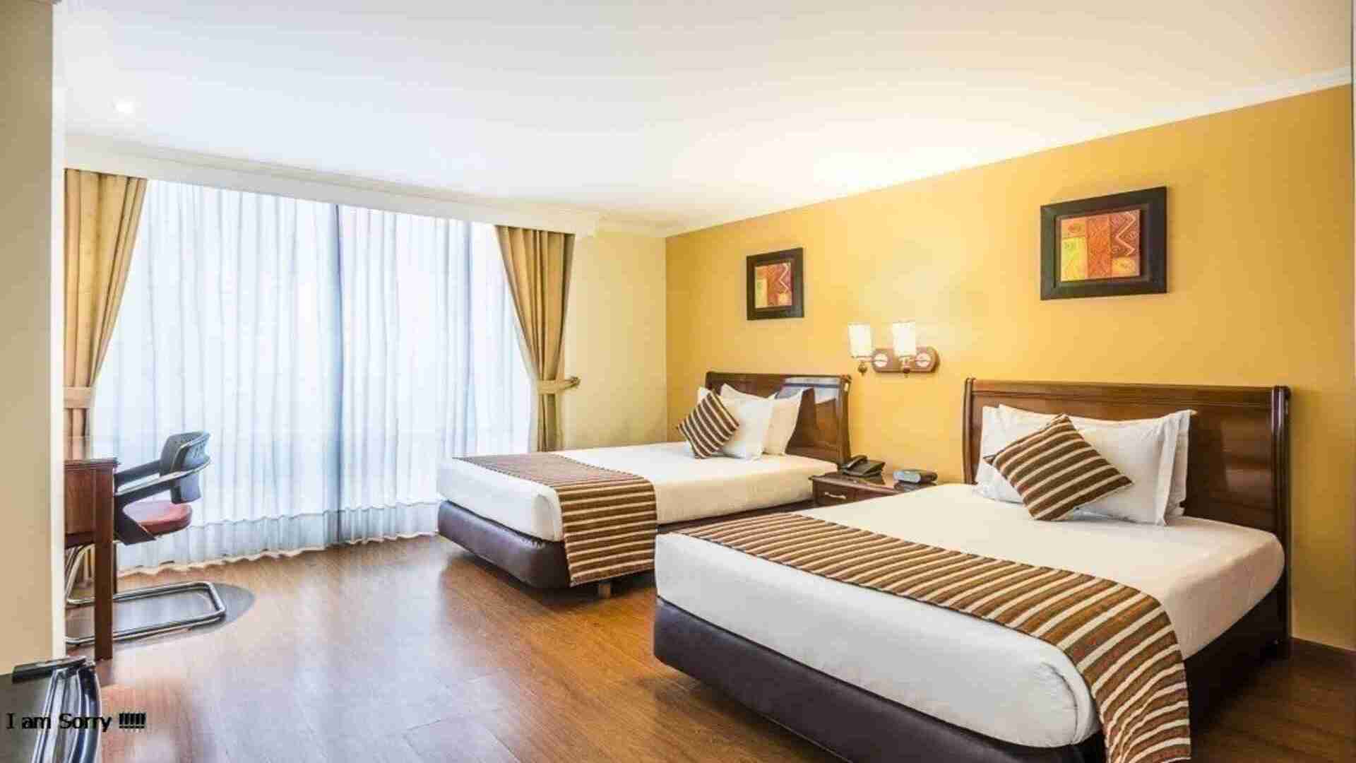 Un dormitorio doble con dos camas individuales, cada una con un camino de cama a rayas. La habitación es espaciosa, luminosa y cuenta con un escritorio.