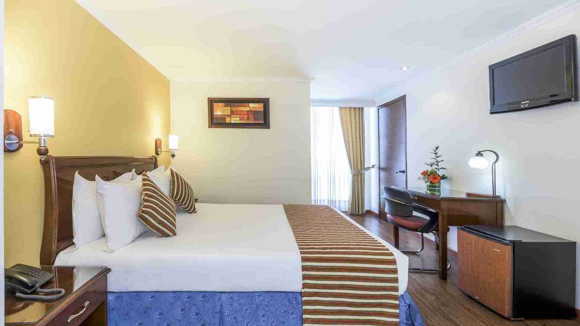 Un dormitorio de hotel con cama king, escritorio, minibar y televisor. La decoración es clásica con tonos cálidos y detalles funcionales.