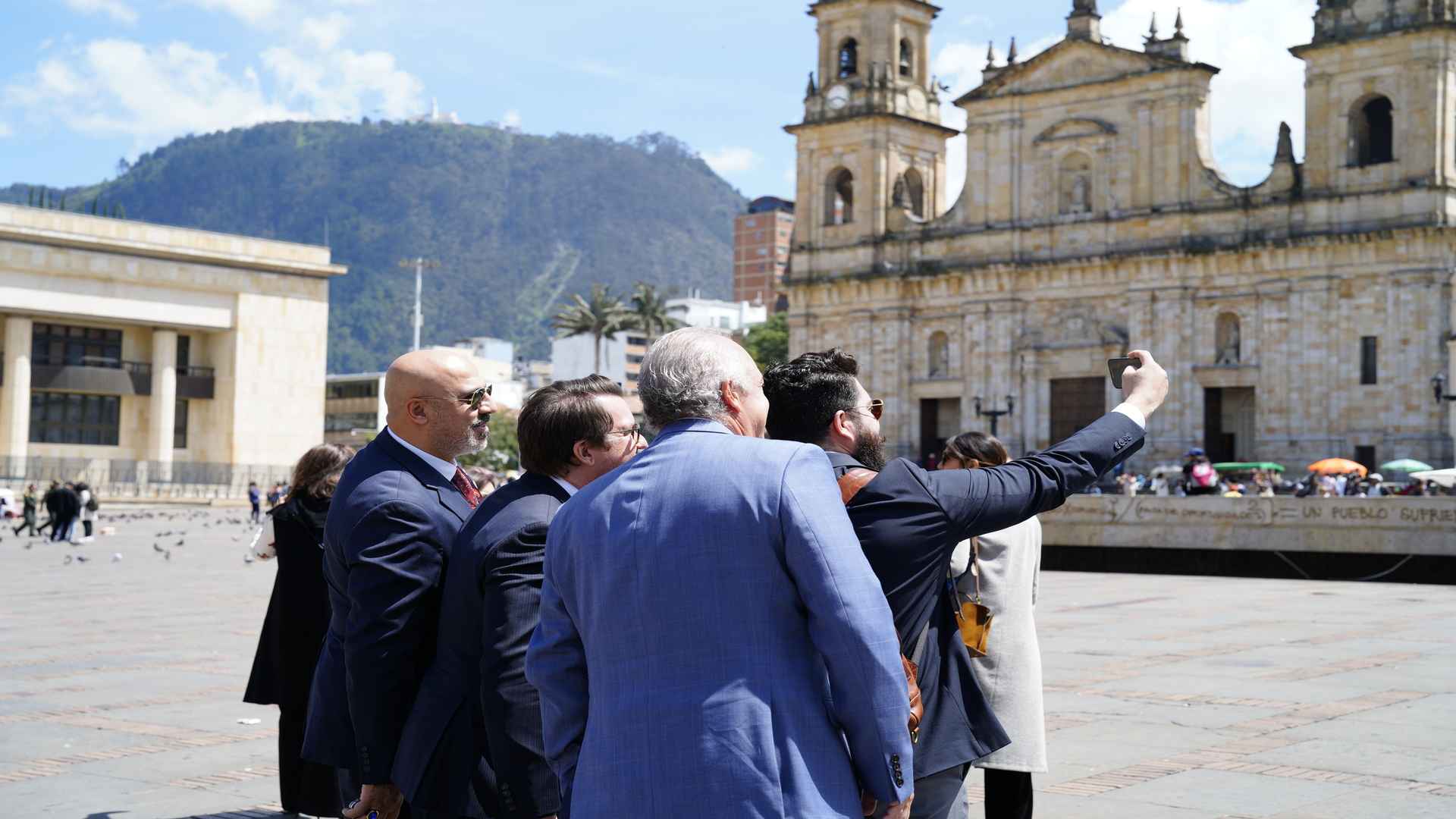 Varias personas de espaldas, una de ellas tomando una selfie, con una iglesia y la plaza principal de fondo. 