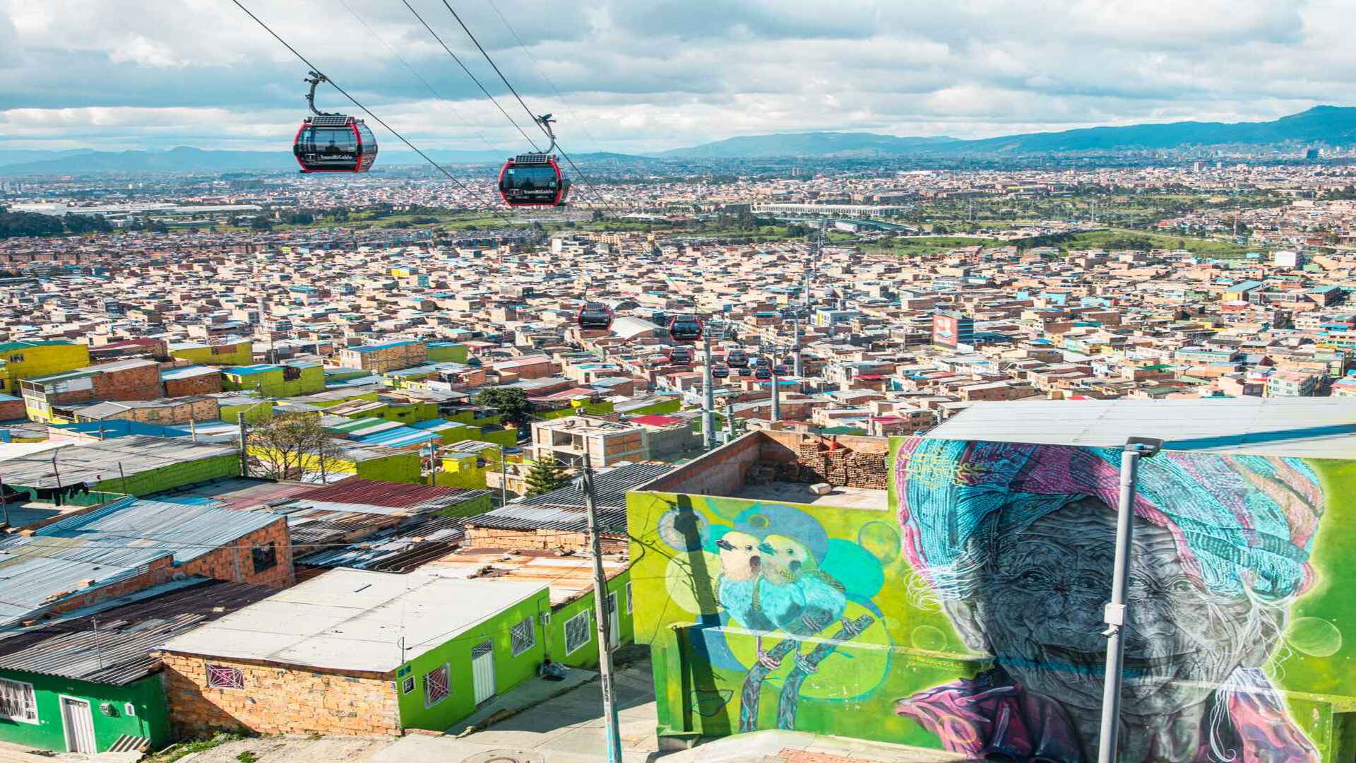 Teleféricos rojos sobrevolando un paisaje urbano denso con casas y un gran mural artístico en Bogotá. 