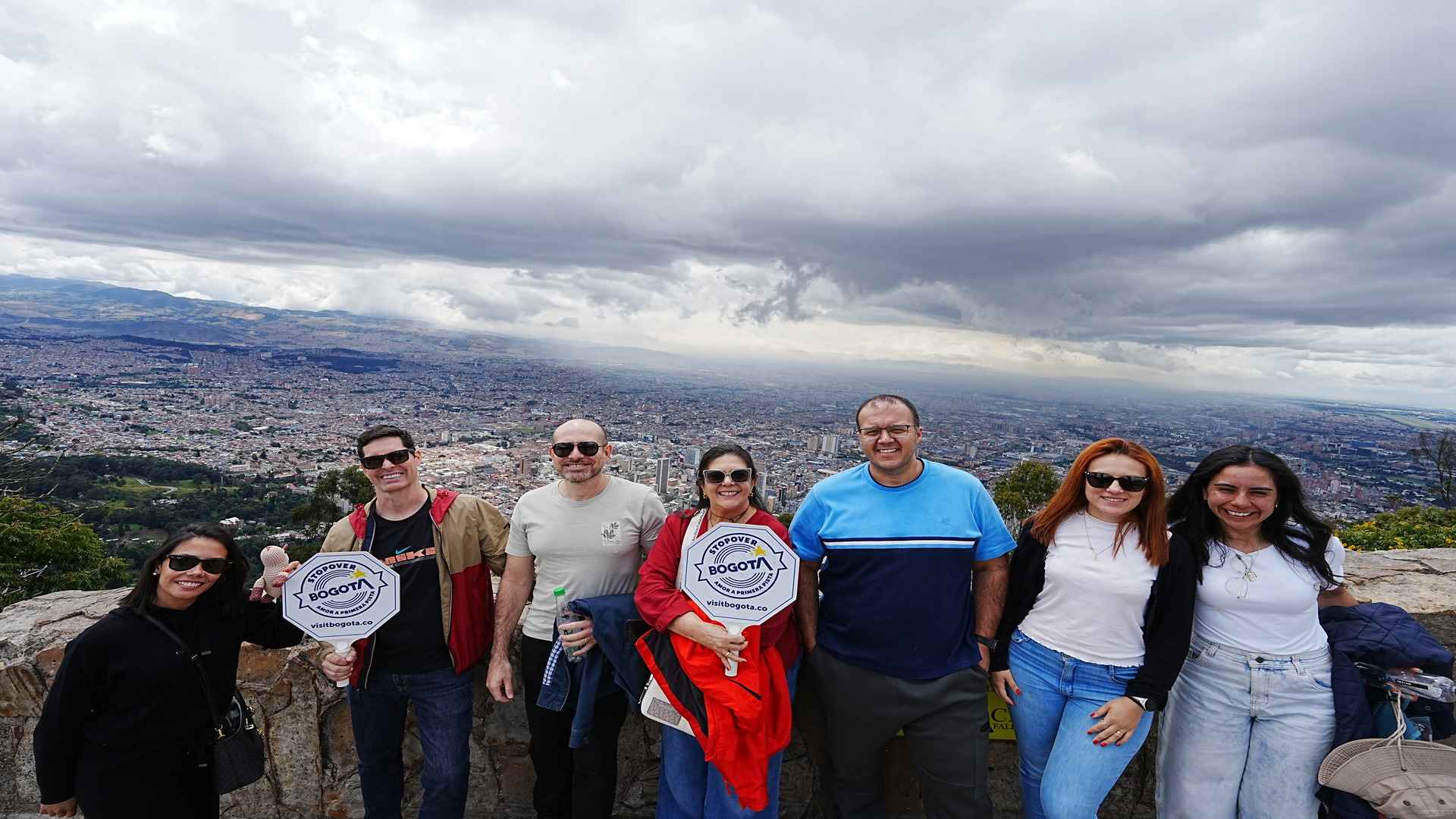  Un grupo de siete personas posando con letreros de "Bogotá" en un mirador con vistas panorámicas de la ciudad.