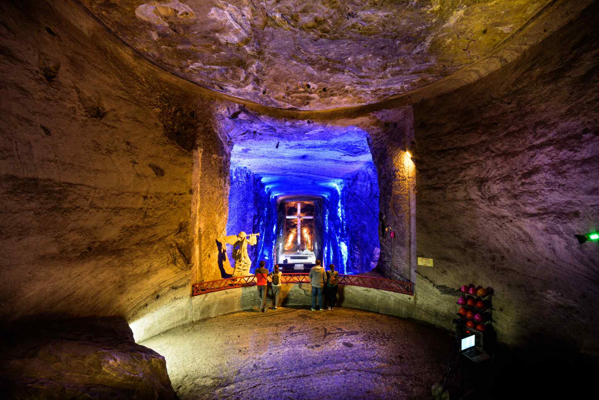 Catedral de Sal paredes de caverna color gris con fondo azul, un ángel y una cruz de concreto. Dos parejas de personas miran al interior de la caverna.