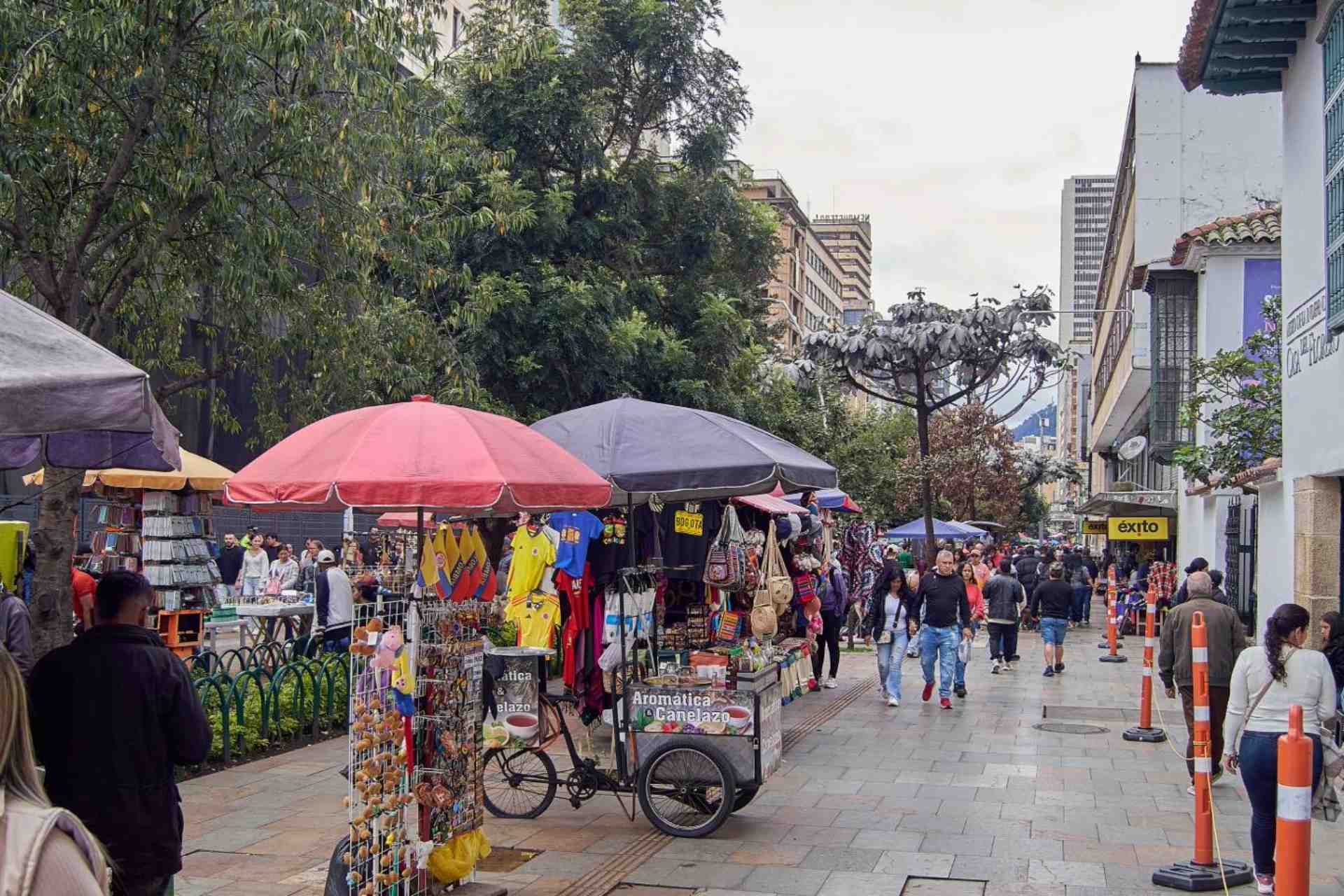 Avenida carrera séptima repleta de vendedores ambulantes con coloridas carpas repletas de souvenirs de Colombia. Decenas de personas camina alrededor y se ven algunos árboles frondosos y al fondo edificios altos.