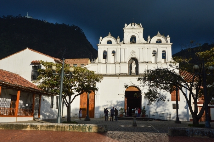 Parroquia Nuestra Señora De Las aguas, fachada de atardecer, cielo gris. Lo rodean en la plaza dos árboles a lado y lado y al fondo los cerros orientales con Monserrate. Hay personas en la puerta.