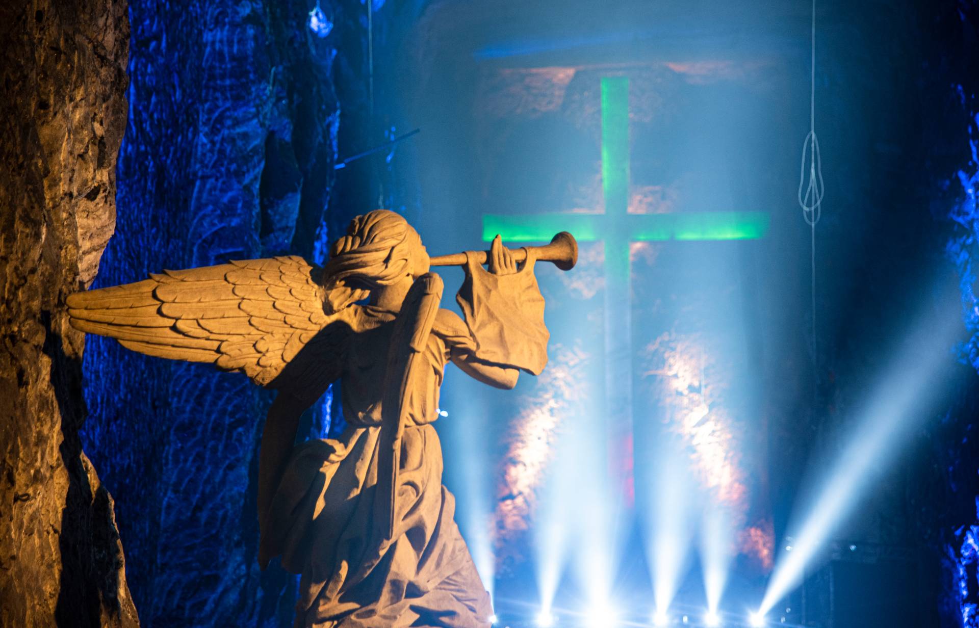  Primer plano de ángel de concreto blanco que toca trompeta mirando hacia el fondo azul de la caverna donde yace una cruz de concreto brillante.