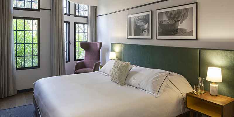 Hotel HAB interior habitación de paredes blancas con cuadros y una cama queen con tendidos blancos y dos mesas de noche con pantallas encendidas. Una sostiene una botella de agua natural y un vaso. Al lado de la cama una poltrona violeta y al fondo seis ventanas grandes con cortinas abiertas.