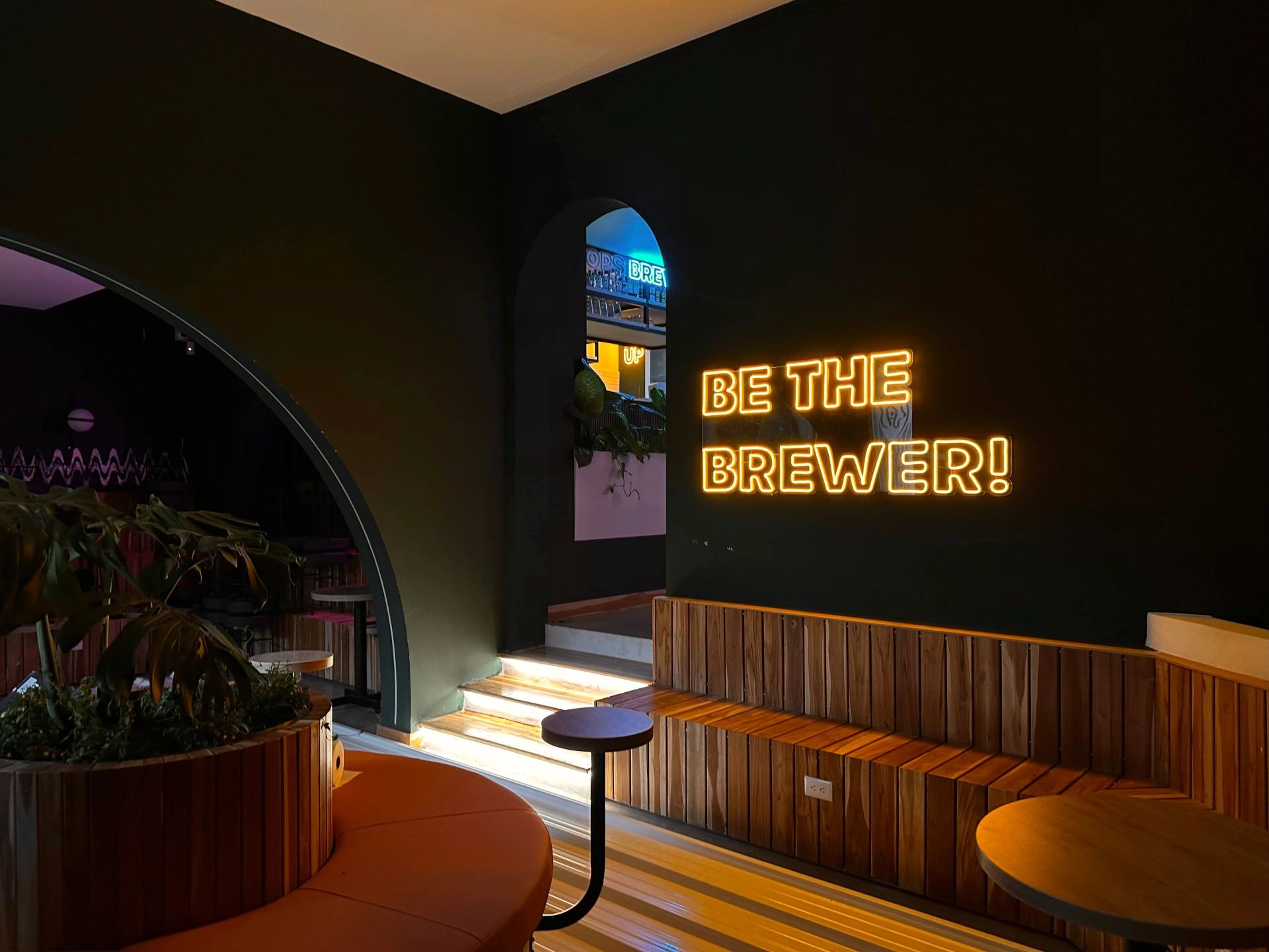 Hanna Hops salón oscuro con paredes pintadas de negro y un aviso de neón en letra en amarilo que dice "Be the Brewer!". Hay una matera al centro rodeada de una silla circular con cojinería.