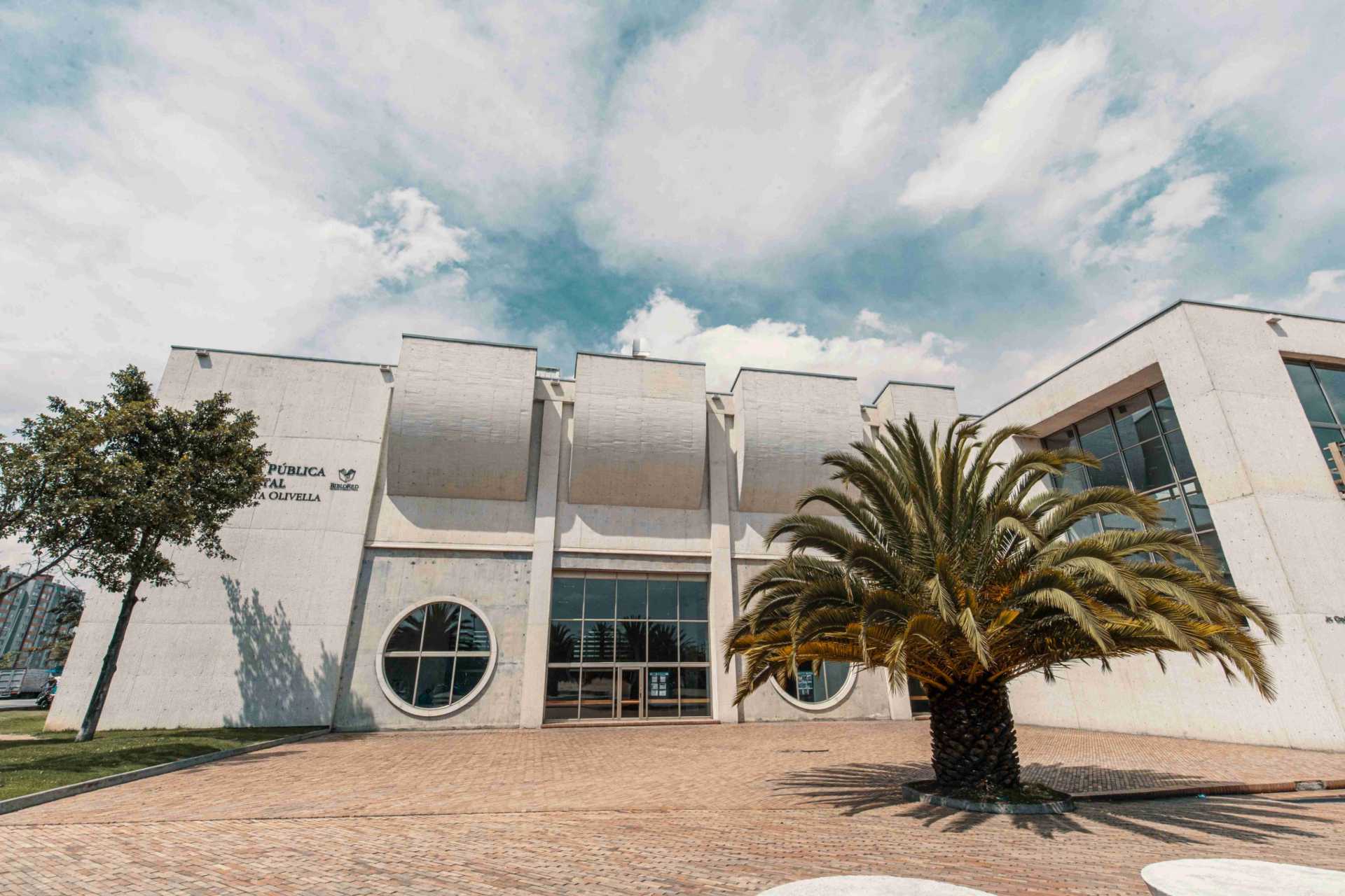 Biblioteca El Tintal fachada de día paredes en concreto blanco con grandes ventanales circulares y cuadrados y gran puerta cuadrada en vidrio. Al frente a la derecha hay una palma de mediano tamaño y a la izquierda un árbol frondoso. El cielo es azul con nubes blancas.