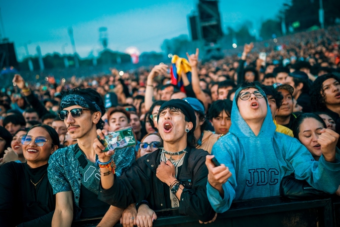 Grupo de personas en concierto al aire libre al atardecer cantando. Sobresalen 5 jóvenes con gorros y balacas y al fondo en la multitud una bandera de Colombia.