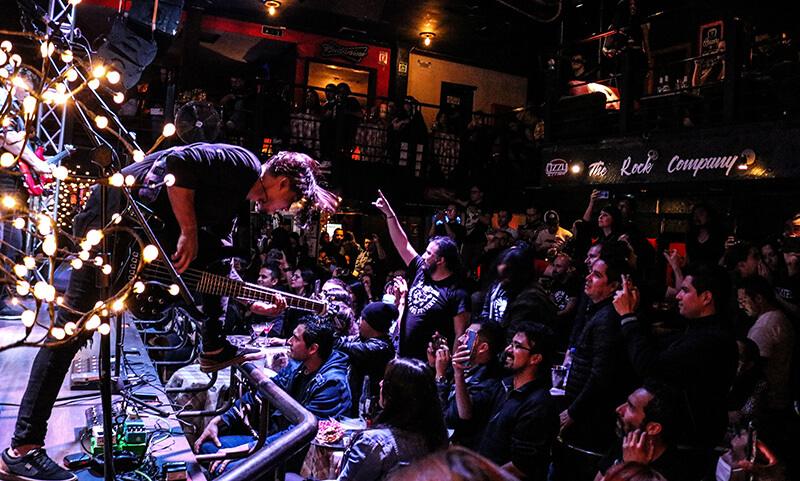 Bar de rock con un guitarrista en el escenario. El lugar está lleno con público de pie, vestido de negro al frente de la tarima. El lugar cuenta  con un segundo piso con personas sentadas.