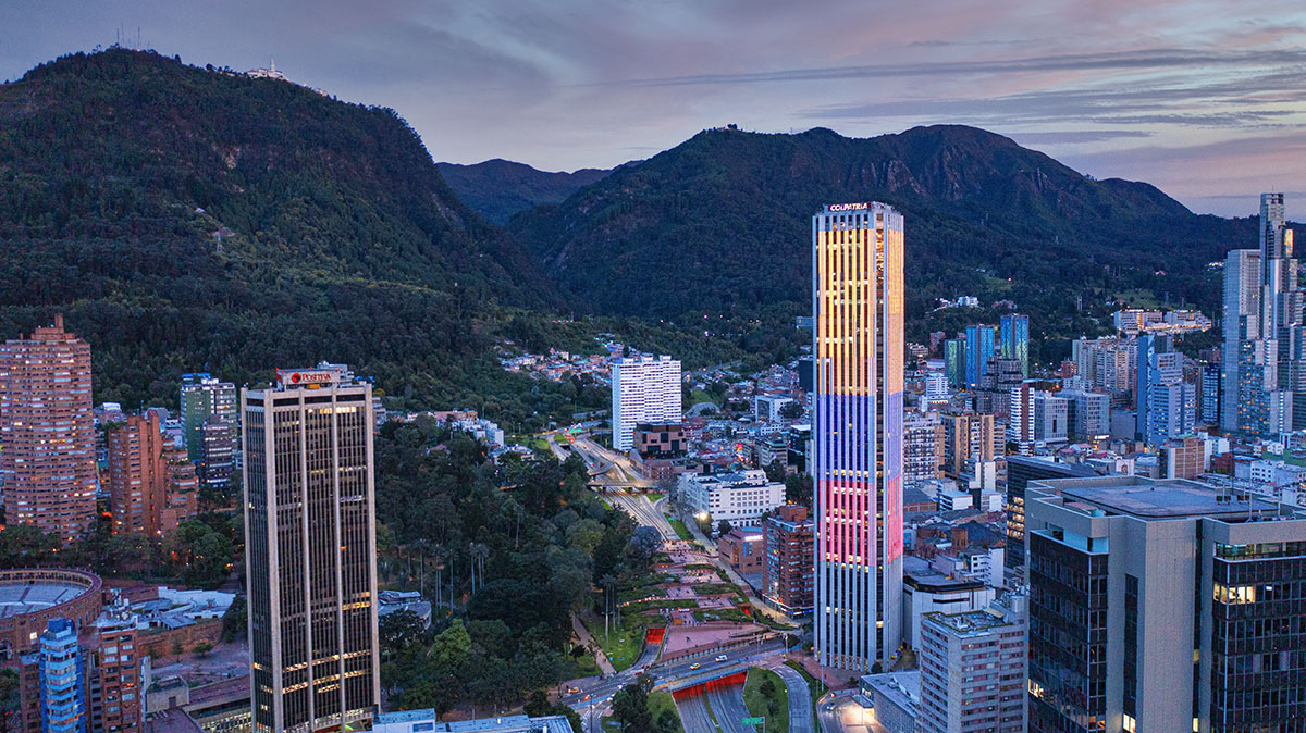 Panorámica del oriente de la ciudad en atardecer con la gigante y colorida Torre Colpatria, edificios altos y Cerros Orientales con montañas altas y verdes.